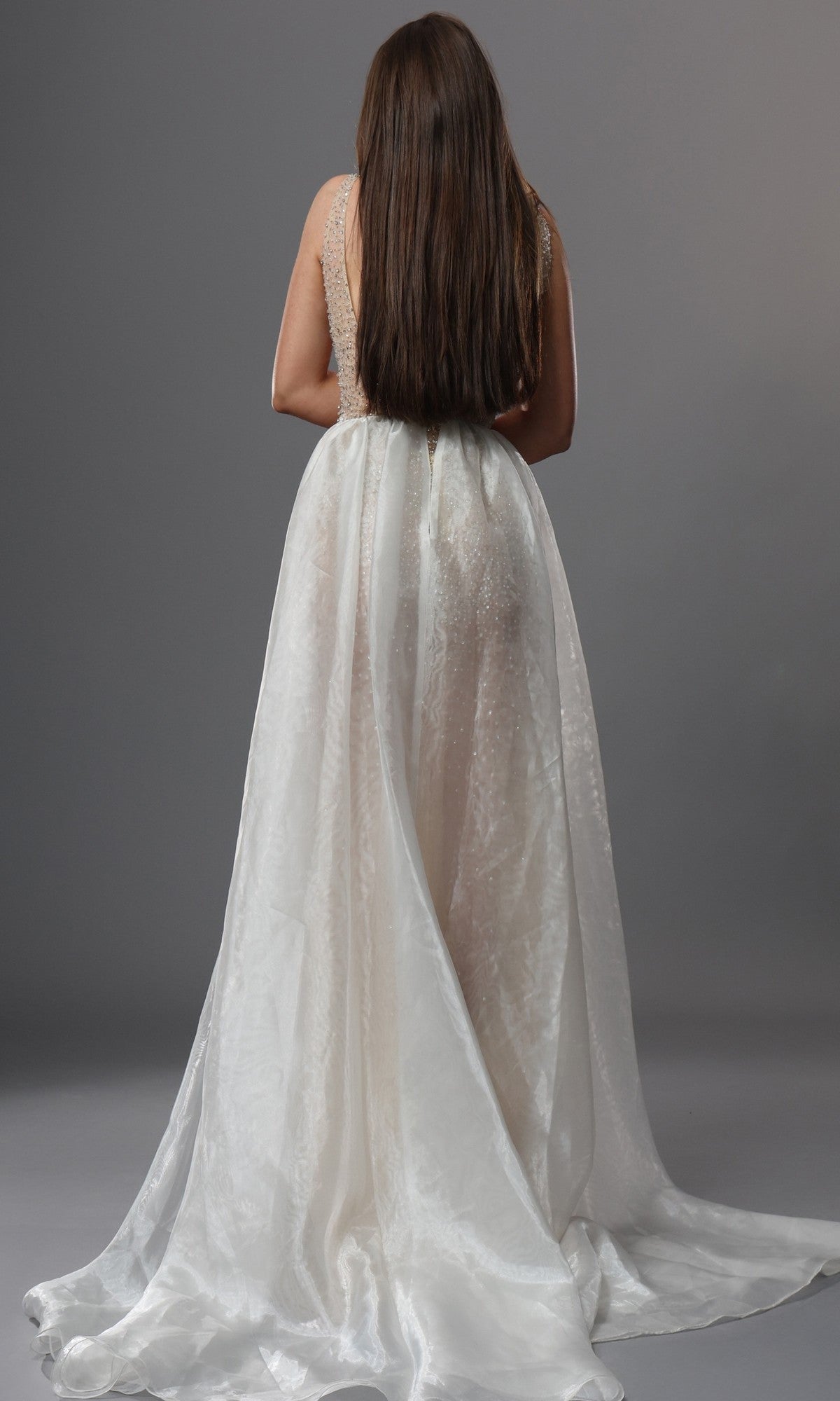 A woman with long brown hair stands facing away, displaying the flowing train of the Jovani 39043 long formal dress against a plain gray background.